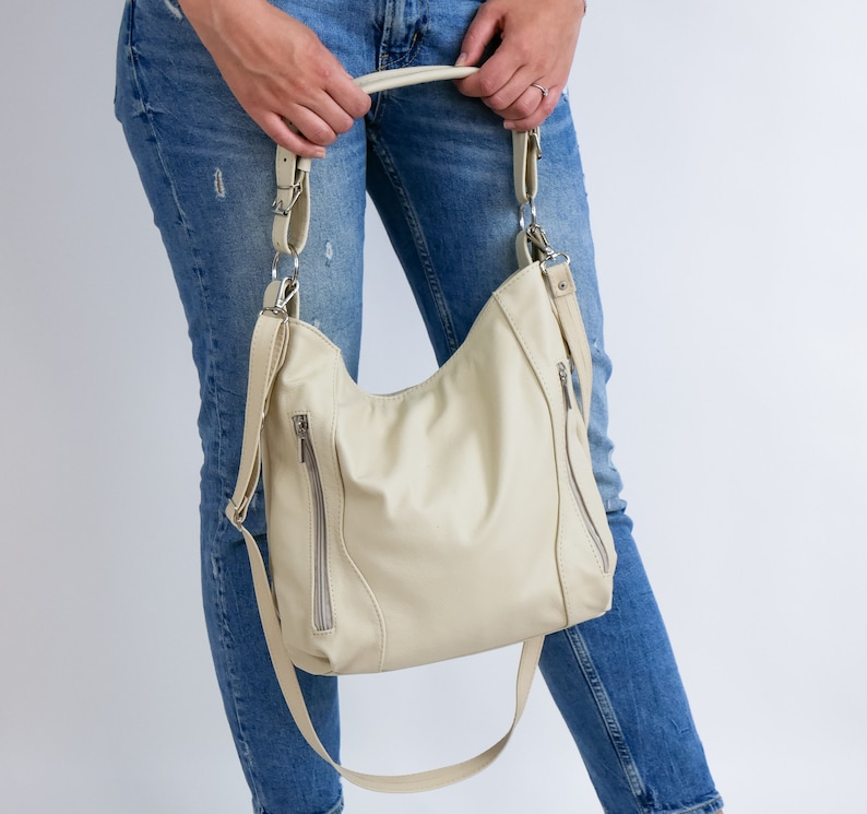 Peut inclure: Un sac &agrave; main en cuir de couleur cr&egrave;me avec des ferrures argent&eacute;es et une fermeture &agrave; glissi&egrave;re. Le sac est port&eacute; par une personne v&ecirc;tue d'un jean bleu.