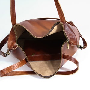May include: A brown leather tote bag with a top zipper closure, two shoulder straps, and antique brass-toned hardware. The interior is lined with a light beige fabric and features a single zippered pocket on the back wall of the bag.