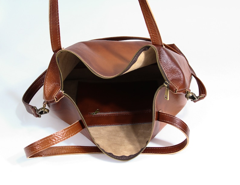 May include: A brown leather tote bag with a top zipper closure, two shoulder straps, and antique brass-toned hardware. The interior is lined with a light beige fabric and features a single zippered pocket on the back wall of the bag.