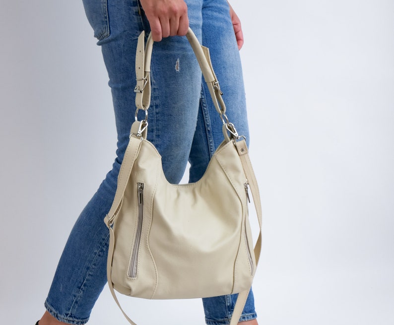 Peut inclure: Un sac &agrave; main en cuir de couleur cr&egrave;me avec une longue bandouli&egrave;re et des ferrures argent&eacute;es. Le sac a une poche zipp&eacute;e sur le devant et une fermeture &agrave; rabat.