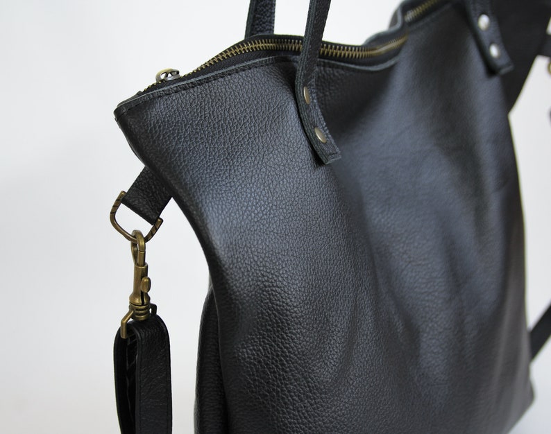 May include: A black pebbled leather tote bag featuring a top zipper closure with brass-toned hardware. The bag includes a detachable shoulder strap connected by a brass swivel clasp and reinforced with metal rivets at the strap attachment points.