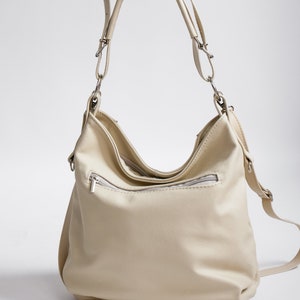 Peut inclure: Un sac &agrave; main en cuir de couleur cr&egrave;me avec une longue bandouli&egrave;re et une poche zipp&eacute;e sur le devant.