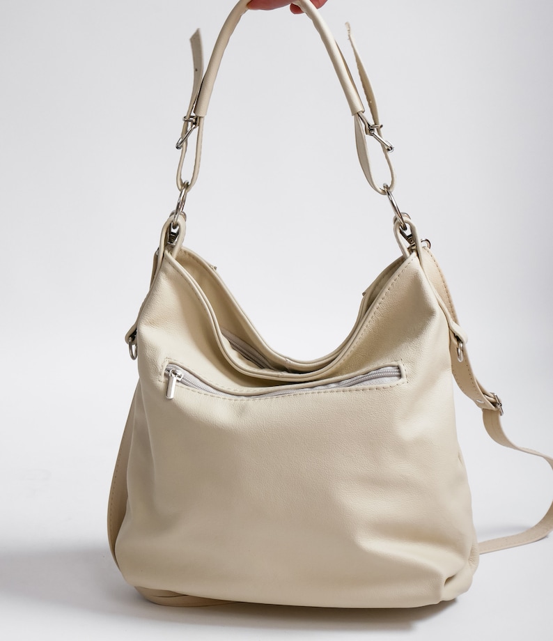 Peut inclure: Un sac &agrave; main en cuir de couleur cr&egrave;me avec une longue bandouli&egrave;re et une poche zipp&eacute;e sur le devant.