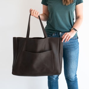 May include: A large, dark brown leather tote bag with a front pocket and long handles. The bag is held up by a person. The bag is designed for carrying items.
