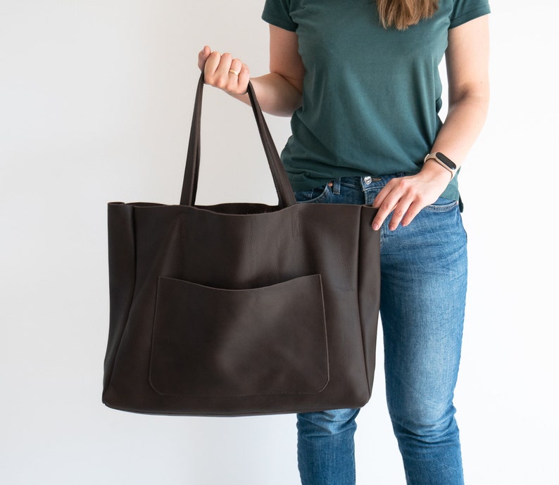 May include: A large, dark brown leather tote bag with a front pocket and long handles. The bag is held up by a person. The bag is designed for carrying items.