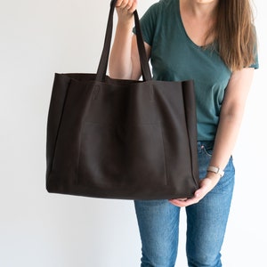 May include: A large, dark brown tote bag with a rectangular shape and two long handles. The bag has a pocket on the front and is held up by a person. The bag is made of a textured material.