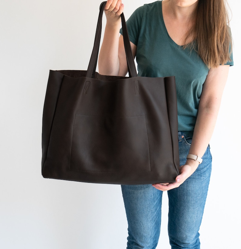 May include: A large, dark brown tote bag with a rectangular shape and two long handles. The bag has a pocket on the front and is held up by a person. The bag is made of a textured material.