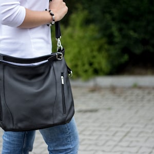 May include: A black leather shoulder bag with a zippered pocket and a silver buckle. The bag is worn over the shoulder of a person wearing a white shirt and blue jeans.