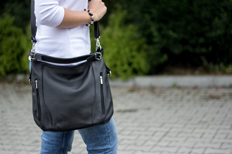 May include: A black leather shoulder bag with a zippered pocket and a silver buckle. The bag is worn over the shoulder of a person wearing a white shirt and blue jeans.