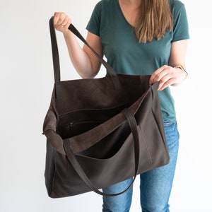 May include: A large, open-top brown leather tote bag with long shoulder straps. The bag has an interior pocket and a contrasting suede-like lining. The bag is held open, showcasing its spacious interior and design.