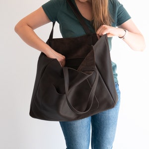 May include: A dark brown leather tote bag with a shoulder strap and exterior pocket. The bag is open, revealing the interior. The tote is being held by a person wearing a green shirt and blue jeans.
