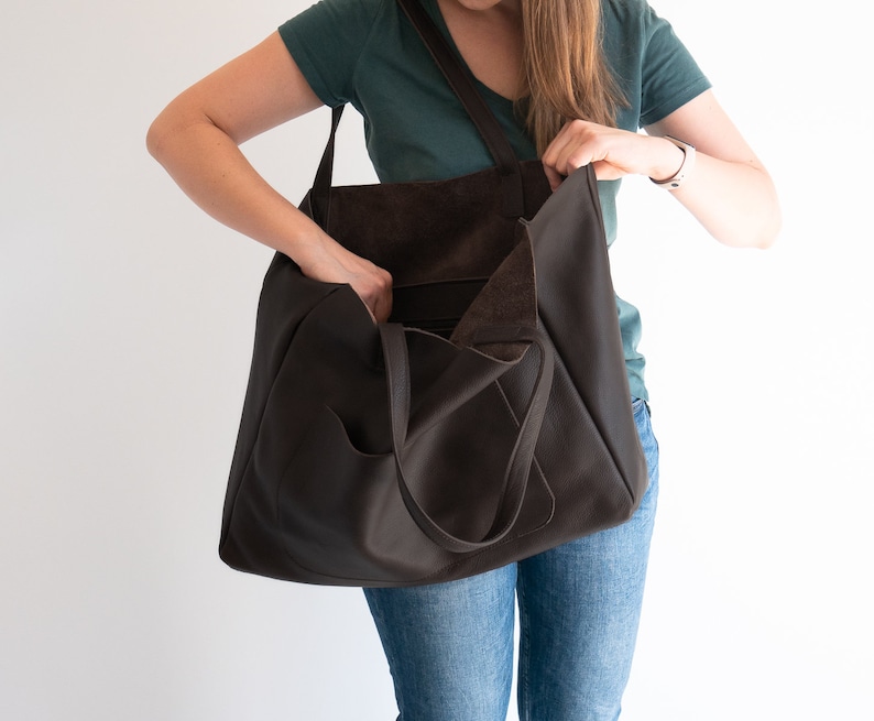 May include: A dark brown leather tote bag with a shoulder strap and exterior pocket. The bag is open, revealing the interior. The tote is being held by a person wearing a green shirt and blue jeans.