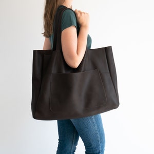 May include: A large, dark brown tote bag with a front pocket and long shoulder straps. The bag is made of a textured material and is being carried by a person. The bag is a practical accessory for carrying items.