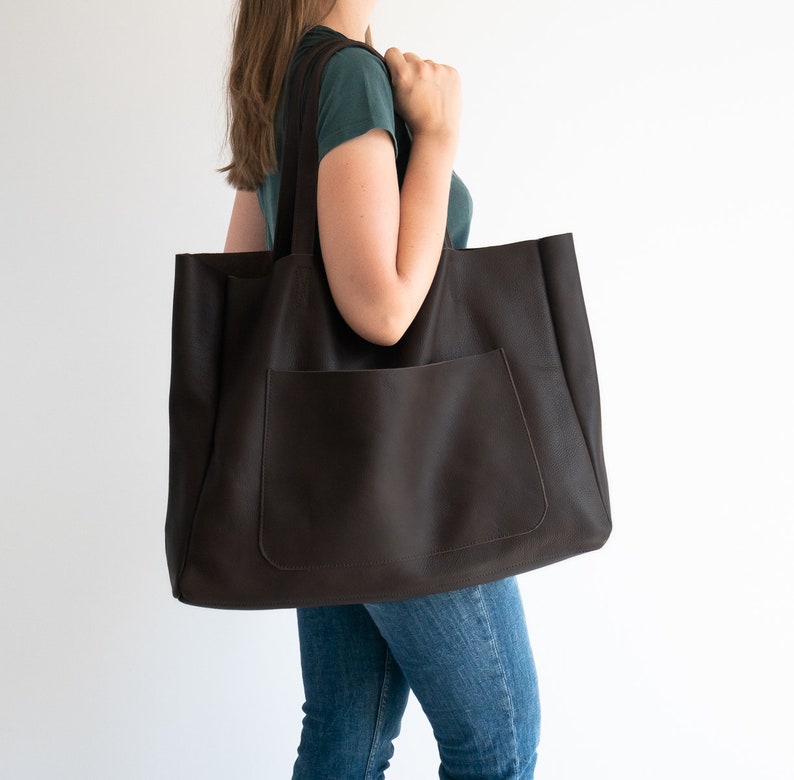 May include: A large, dark brown tote bag with a front pocket and long shoulder straps. The bag is made of a textured material and is being carried by a person. The bag is a practical accessory for carrying items.