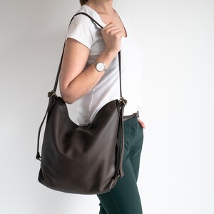 May include: A dark brown hobo-style shoulder bag with a slouchy silhouette, featuring thin adjustable straps, antique brass-toned metal hardware, and a smooth, matte finish. The bag is worn over the shoulder, showcasing its flexible structure and simple design.