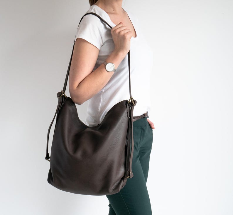 May include: A dark brown hobo-style shoulder bag with a slouchy silhouette, featuring thin adjustable straps, antique brass-toned metal hardware, and a smooth, matte finish. The bag is worn over the shoulder, showcasing its flexible structure and simple design.
