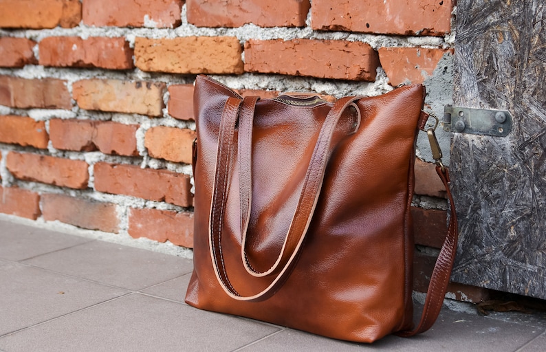 May include: A brown leather tote bag with a textured surface, featuring two long shoulder straps and a top zipper closure. The bag has a rectangular shape with rounded corners and includes a side attachment point for an additional strap.