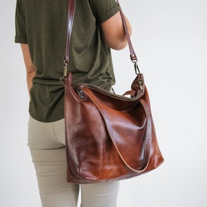 May include: A large, deep brown leather shoulder bag with a textured surface, featuring a top zipper closure, two short handles, and a long, adjustable crossbody strap attached with antique brass-toned metal hardware clips.