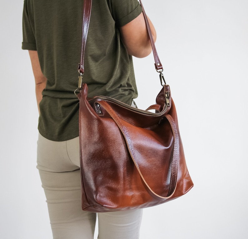 May include: A large, deep brown leather shoulder bag with a textured surface, featuring a top zipper closure, two short handles, and a long, adjustable crossbody strap attached with antique brass-toned metal hardware clips.