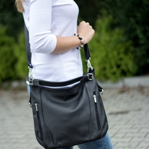 May include: A black leather shoulder bag with silver hardware and a zippered pocket. The bag is worn over the shoulder of a person wearing a white shirt and blue jeans.