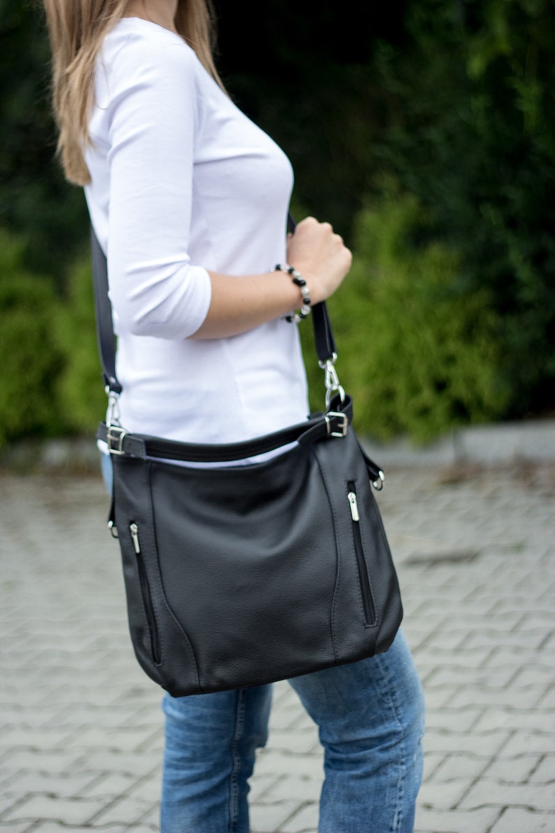 May include: A black leather shoulder bag with silver hardware and a zippered pocket. The bag is worn over the shoulder of a person wearing a white shirt and blue jeans.