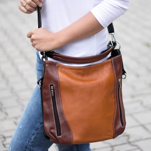 Puede incluir: Un bolso de hombro de cuero marr&oacute;n y beige con una correa negra. El bolso tiene dos bolsillos con cremallera en la parte delantera.