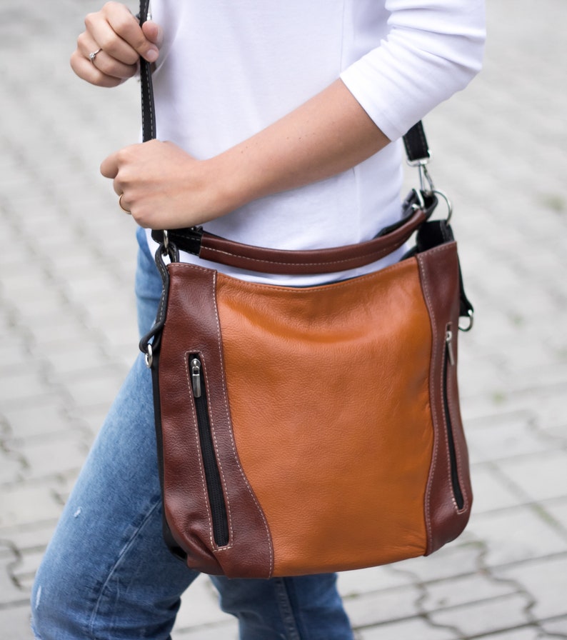 Puede incluir: Un bolso de hombro de cuero marr&oacute;n y beige con una correa negra. El bolso tiene dos bolsillos con cremallera en la parte delantera.