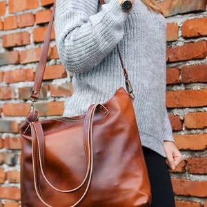 May include: A large, cognac-brown leather tote bag featuring a smooth, polished finish, two top handles, and a detachable shoulder strap with antique brass-toned metal hardware, including a top zipper closure and visible stitching details.