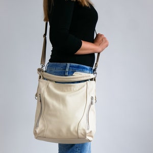 Peut inclure: Un sac &agrave; bandouli&egrave;re en cuir cr&egrave;me avec des d&eacute;tails argent&eacute;s et des accents de fermeture &eacute;clair. Le sac est port&eacute; sur l'&eacute;paule d'une personne portant un jean bleu.