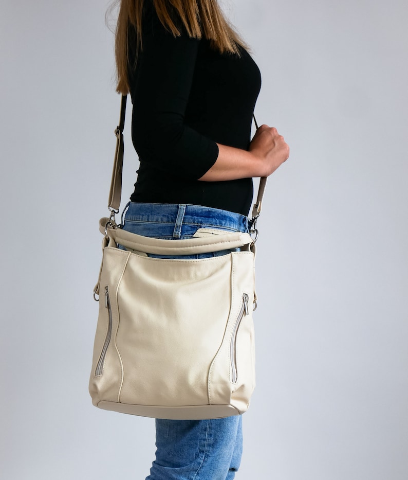 Peut inclure: Un sac &agrave; bandouli&egrave;re en cuir cr&egrave;me avec des d&eacute;tails argent&eacute;s et des accents de fermeture &eacute;clair. Le sac est port&eacute; sur l'&eacute;paule d'une personne portant un jean bleu.