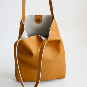 May include: A mustard yellow leather tote bag with white stitching and a white leather lining. The bag has a single top handle and a long shoulder strap.
