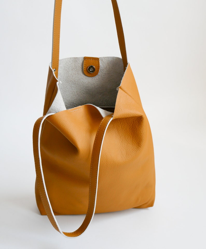 May include: A mustard yellow leather tote bag with white stitching and a white leather lining. The bag has a single top handle and a long shoulder strap.