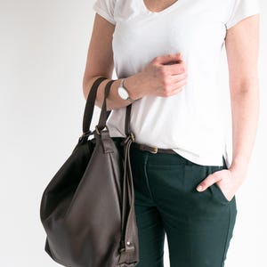 May include: A dark brown leather hobo bag with two top handles and brass-toned hardware. The bag features a slouchy, unstructured silhouette with a pebbled texture, hanging from the shoulder with visible stitching and metal ring attachments.