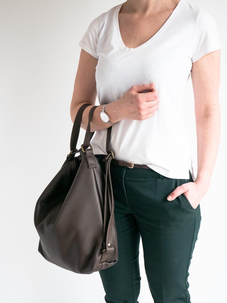 May include: A dark brown leather hobo bag with two top handles and brass-toned hardware. The bag features a slouchy, unstructured silhouette with a pebbled texture, hanging from the shoulder with visible stitching and metal ring attachments.