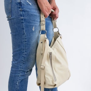Peut inclure: Un sac &agrave; bandouli&egrave;re en cuir de couleur cr&egrave;me avec une poche zipp&eacute;e et une longue bandouli&egrave;re. Le sac est port&eacute; sur l'&eacute;paule d'une personne portant un jean bleu.