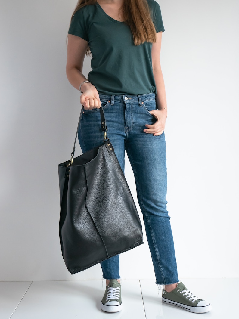 K&ouml;nnte beinhalten: Eine Frau tr&auml;gt ein t&uuml;rkisfarbenes T-Shirt und eine blaue Jeans. Sie h&auml;lt eine gro&szlig;e schwarze Ledertasche mit einem langen Riemen.
