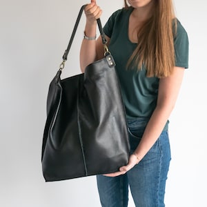 K&ouml;nnte beinhalten: Eine gro&szlig;e schwarze Ledertasche mit einem langen Trageriemen. Die Tasche wird von einer Person getragen, die ein gr&uuml;nes Shirt und eine blaue Jeans tr&auml;gt.