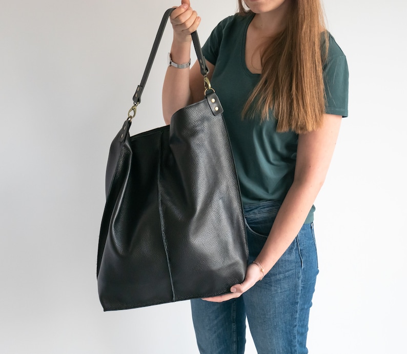 K&ouml;nnte beinhalten: Eine gro&szlig;e schwarze Ledertasche mit einem langen Trageriemen. Die Tasche wird von einer Person getragen, die ein gr&uuml;nes Shirt und eine blaue Jeans tr&auml;gt.