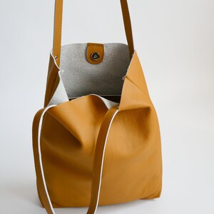 May include: A mustard yellow leather tote bag with white trim and a single snap closure. The bag has a long strap that can be worn on the shoulder or across the body.