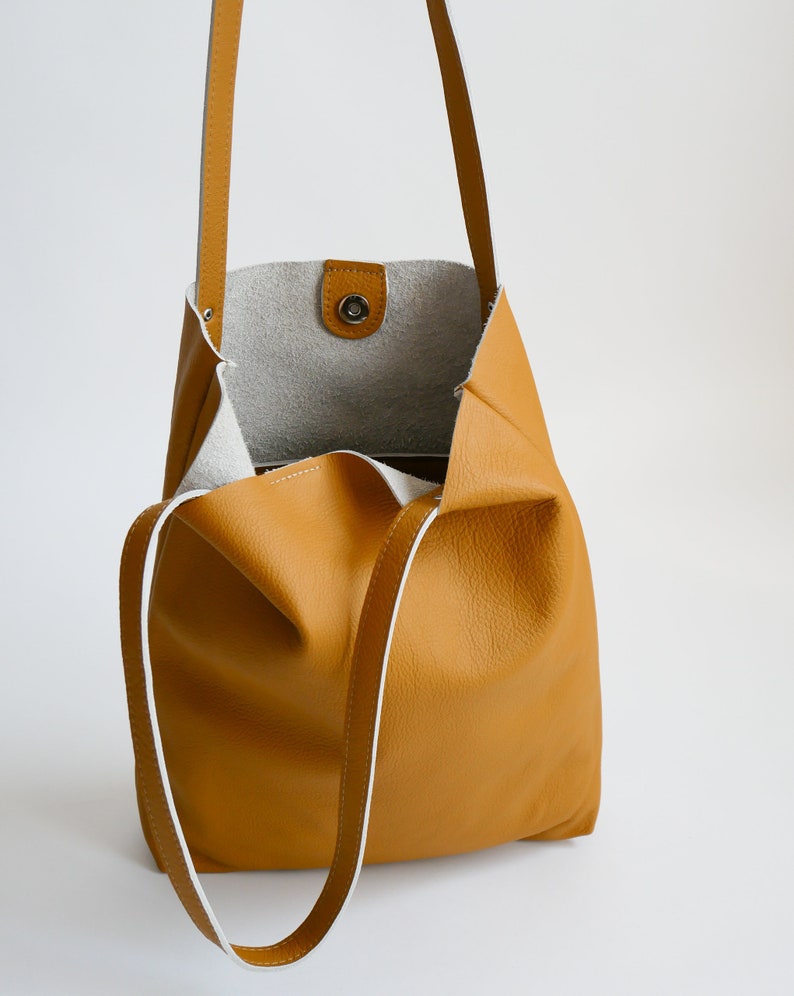 May include: A mustard yellow leather tote bag with white trim and a single snap closure. The bag has a long strap that can be worn on the shoulder or across the body.