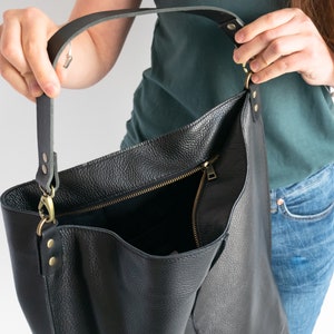 K&ouml;nnte beinhalten: Eine schwarze Ledertasche mit Rei&szlig;verschluss und einem langen, verstellbaren Trageriemen. Die Tasche ist offen und das Innere ist sichtbar.