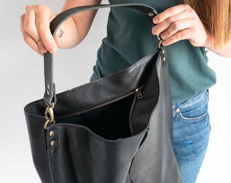 K&ouml;nnte beinhalten: Eine schwarze Ledertasche mit Rei&szlig;verschluss und einem langen, verstellbaren Trageriemen. Die Tasche ist offen und das Innere ist sichtbar.