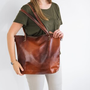 May include: A large, cognac-brown leather tote bag with a smooth, slightly distressed finish. The bag features two long shoulder straps attached with metal rivets and a top zipper closure. The leather has a rich, deep brown color variation.