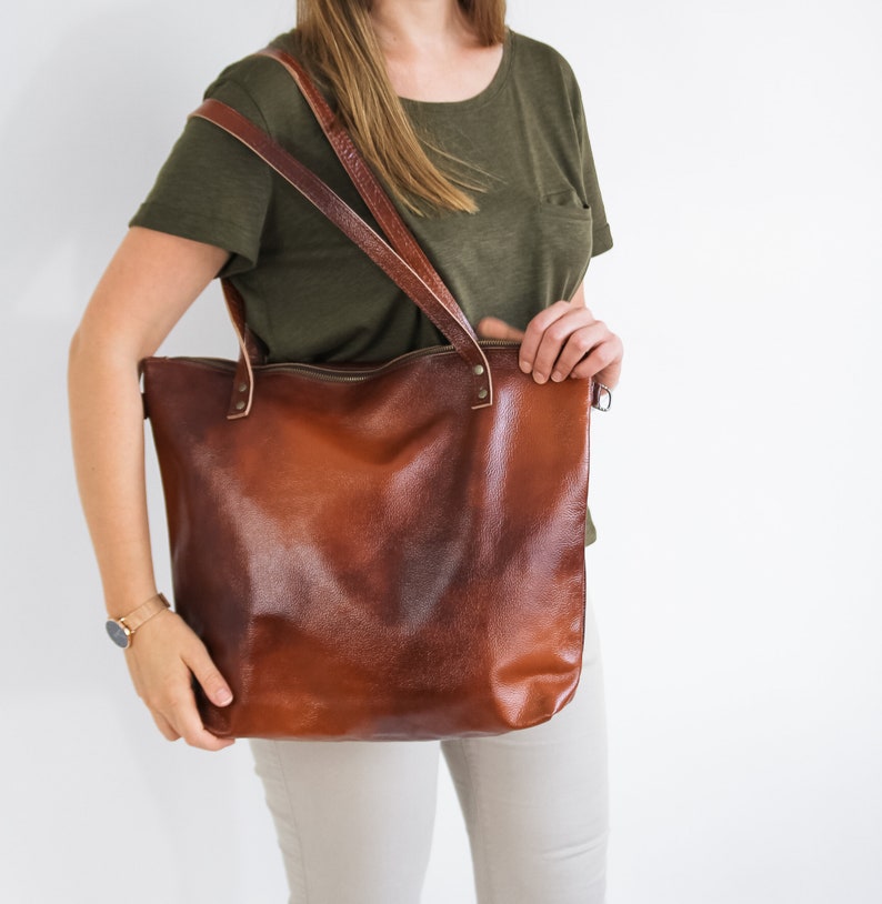 May include: A large, cognac-brown leather tote bag with a smooth, slightly distressed finish. The bag features two long shoulder straps attached with metal rivets and a top zipper closure. The leather has a rich, deep brown color variation.