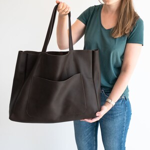 May include: A large, dark brown leather tote bag with a front pocket and long handles. The bag is held up by a person. The bag is designed for carrying items and has a simple, classic design.