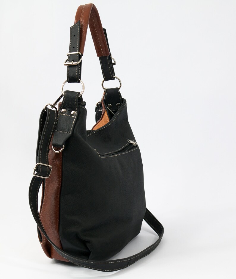 Puede incluir: Un bolso de mano de cuero negro con ribete de cuero marr&oacute;n y una correa larga para el hombro. El bolso tiene un bolsillo con cremallera en la parte delantera.