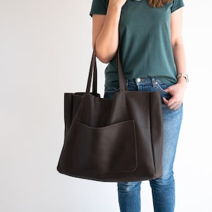 May include: A large, dark brown tote bag with a front pocket and long handles. The bag is made of a textured material and is being held by a person wearing a green shirt and blue jeans. The bag appears to be empty.