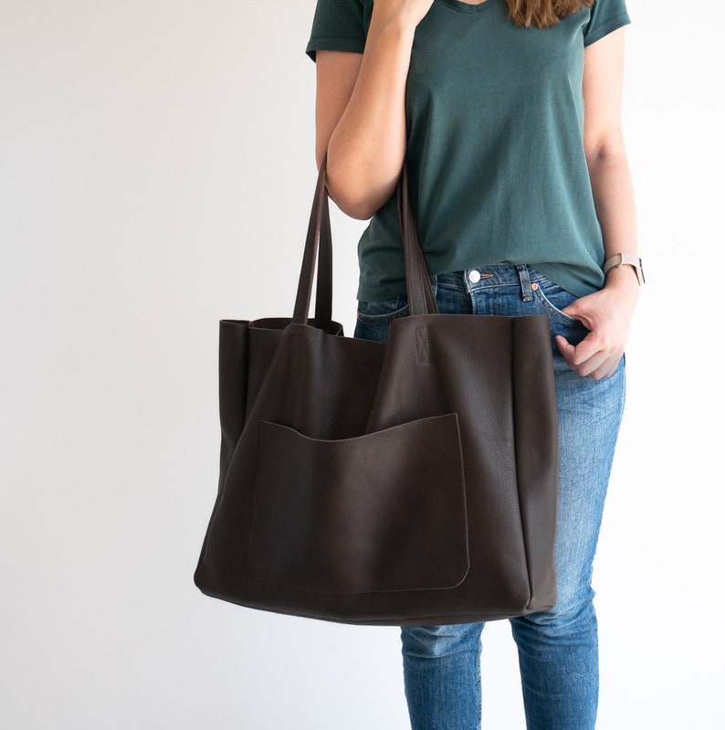 May include: A large, dark brown tote bag with a front pocket and long handles. The bag is made of a textured material and is being held by a person wearing a green shirt and blue jeans. The bag appears to be empty.