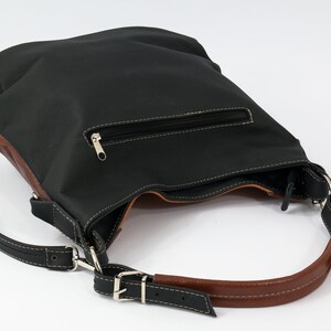 Puede incluir: Un bolso de hombro de cuero negro con ribete de cuero marr&oacute;n. El bolso tiene un bolsillo con cremallera en la parte delantera y una correa larga ajustable.