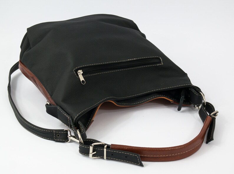 Puede incluir: Un bolso de hombro de cuero negro con ribete de cuero marr&oacute;n. El bolso tiene un bolsillo con cremallera en la parte delantera y una correa larga ajustable.